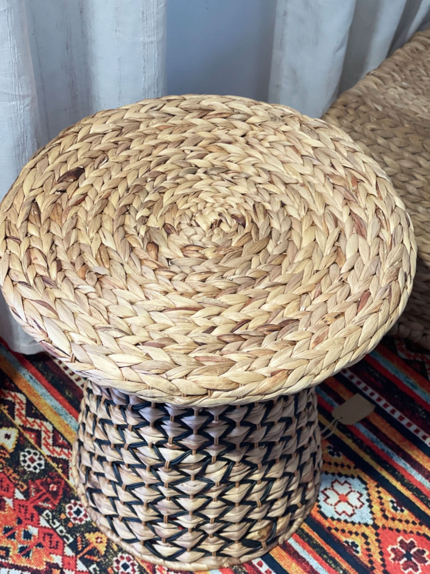 Wabi Sabi Coffee Table Straw: Round woven natural straw top with black and tan braided base on a colorful rug.